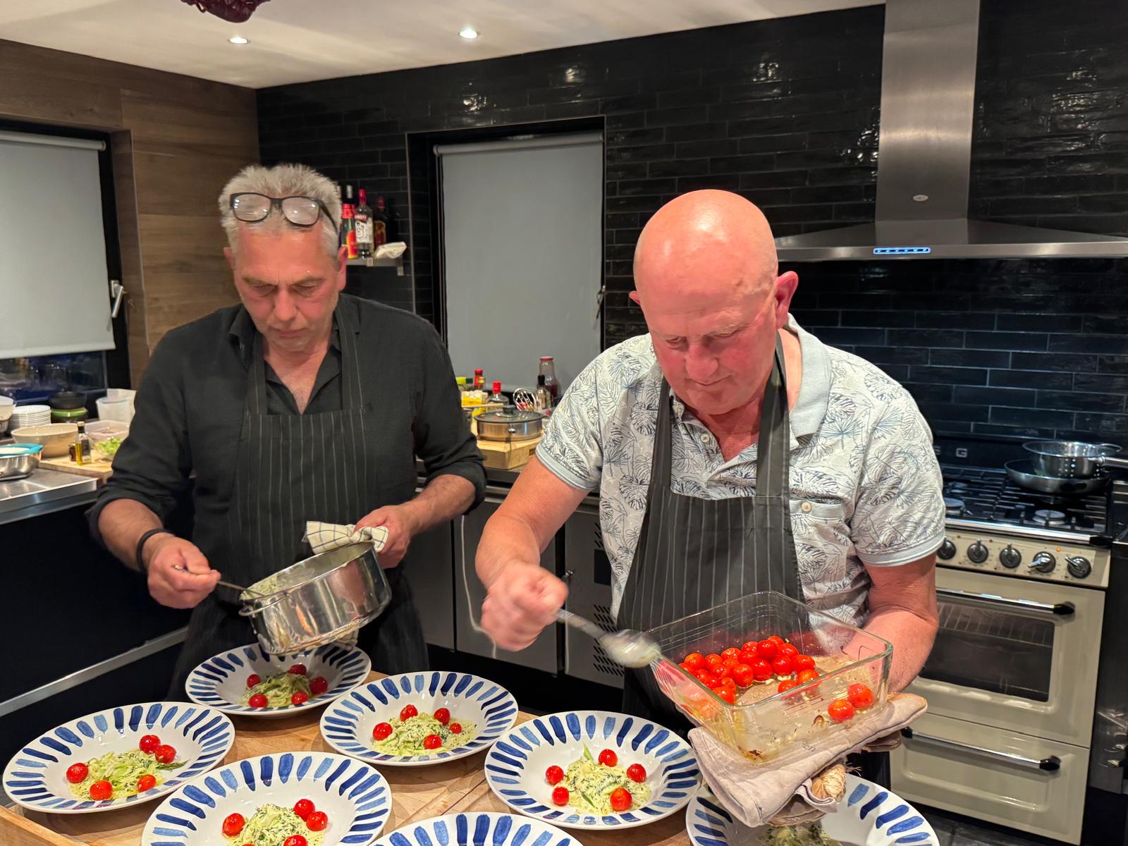 Twee kels werken aan een rij pastaborden, met kerstomaten erover.